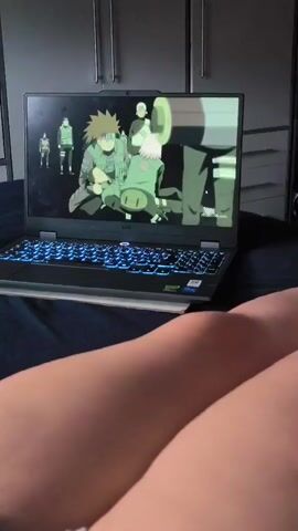 A laptop is open on a dark blue surface, possibly a bed. The laptop screen displays an animated scene. In the foreground, the legs of a person are visible, out of focus. The animation on the laptop screen depicts several characters, seemingly from an anime series. The first scene shows three characters in green outfits and a large white dog-like creature. The second scene shows a single male character with blonde hair, a headband, and a scar. The laptop has blue LED lighting around the keyboard. The background shows closet doors.