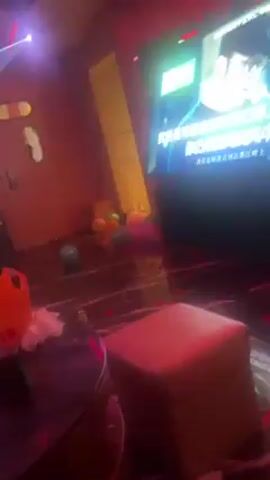 The video shows the interior of a karaoke room. A large screen displays a video of a man singing with subtitles in Chinese. The room is dimly lit with red and white neon lights. There is a small tan ottoman in the foreground and various toys scattered on the floor. The room also features a doorway, an arcade game, and a sofa. Music plays in the background.