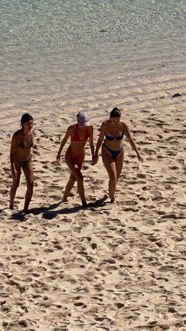 Three women walk parallel to each other on a sandy beach on a sunny day. One woman wears a black bikini with white polka dots, another wears a red bikini and a baseball cap, and the third wears a blue bikini. They are walking away from the viewer and toward the clear water. The sand is textured and dry.
