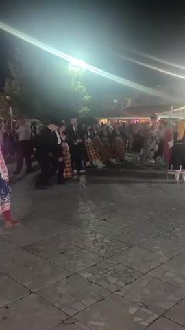 A group of people are dancing in a public square at night. The dancers are wearing traditional clothing, including kilts and skirts with patterns, and some are wearing black suits. There is a crowd of onlookers watching the performance. Bright spotlights illuminate the dancing area, creating strong contrasts between light and shadow. The ground is paved with square tiles. In the background, buildings and some greenery are visible. The overall atmosphere appears festive.