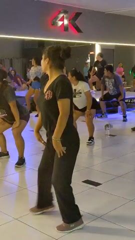 A group of people, predominantly women, are engaged in an energetic indoor fitness class, likely a dance or aerobics session. They are moving in synchronized fashion, following an instructor (partially visible in the foreground initially) within a gym or studio setting. The participants are dressed in athletic attire, including t-shirts, leggings, shorts, and sneakers.

The room features a light-tiled floor and mirrors along one wall, reflecting the class. Artificial lighting illuminates the space, with blue and green spotlights casting colors onto the floor and a disco ball hanging from the ceiling, adding to the dynamic atmosphere. In the background, a large red and black "4K" logo is prominently displayed on the wall, and some workout benches or equipment platforms are visible. The overall scene suggests a vibrant and engaging fitness environment.