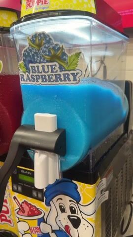The video shows a close-up of a blue raspberry slushie machine in operation. The machine is a multi-compartment unit with a clear plastic container filled with blue, slushy liquid. A black handle is attached to the dispensing spout, which is white. Above the container, a colorful label depicts blueberries and the words "BLUE RASPBERRY". The machine is situated on top of another unit with a yellow and white design, featuring cartoon images. A portion of a red slushie is visible in a cup on this lower unit. The lighting suggests an indoor environment, likely a convenience store or similar establishment. The slushie is actively churning in the machine.