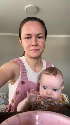 A woman with dark hair pulled back wears a white tank top and pink overalls, holding a baby with blue eyes and fair hair. The woman is smiling as she feeds the baby with a spoon from a pink bowl. The baby is wearing a yellow and white outfit with a banana print. The background shows a plain wall with a light fixture on the ceiling.