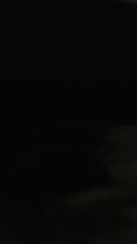 The video shows a nighttime scene through a window. The scene is mostly dark with some blurry lights. The first few seconds show two small, blurry, pale green lights. Then, a long, blurry horizontal streak of green and white lights appears, suggesting movement. Towards the end, there are more blurry lights, some appearing red, moving horizontally across the frame. The overall impression is of passing lights in the darkness, possibly from vehicles on a road or distant buildings.