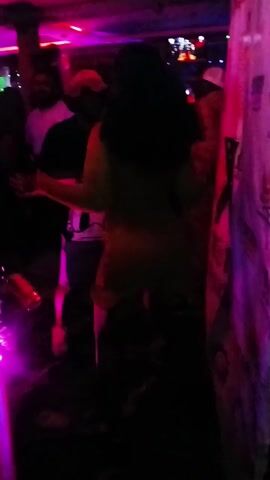 The video shows a dimly lit room, possibly a nightclub, with various people dancing. One person in the foreground is wearing a yellow dress. Another individual wears a black t-shirt with a Donald Duck graphic. A man with a beard is wearing a white t-shirt. The lighting cycles through blue, pink, and red hues. Some of the dancers are partially obscured by another structure.