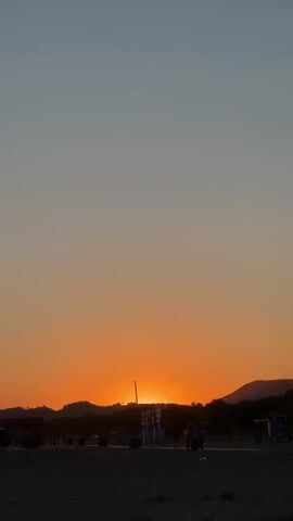 This is a vertical video of a beach at sunset. The sun is setting behind a line of hills, casting an orange glow across the sky. The sand is dark, and there are footprints visible. In the foreground, there is a blanket with a colorful pattern, and a person is lying on it, looking out at the sunset. There are also some beach chairs and umbrellas in the background. The sky is a gradient of blue and orange, with the blue fading to orange towards the horizon.
