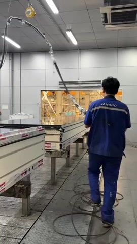 A manufacturing facility is shown in this video, specifically focusing on a work station where glass panes are being assembled into window frames. There is a worker, wearing a blue uniform, using a tool with a cylindrical handle and a flat end to seemingly press or seal the glass into the frame. The frame is white with red markings, sitting on a raised metal stand. The worker is wearing work gloves and appears to be inspecting the window as he works. The environment is a well-lit, clean industrial space with machinery and other materials visible in the background. The facility appears to be engaged in the mass production of windows.