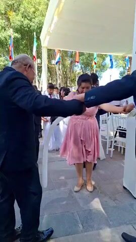 Two older men in dark suits are dancing with a woman in a pink dress at an outdoor event. The woman is holding a bouquet of white flowers. They are under a white awning, and in the background, other people are dressed formally, some in suits and others in dresses.  Flags are hanging in the background. The ground is paved with gray stone. The men are holding the woman's hands and twirling her around. The woman is smiling and looking at the camera.  The atmosphere appears festive.