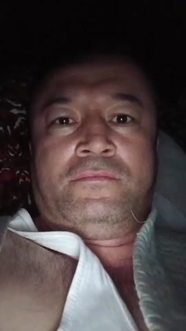 A man is lying down in dim lighting, looking directly at the camera. He is wearing a white shirt and has short dark hair. He briefly shows a peace sign with his fingers. The background includes a dark patterned surface and a light-colored covering.