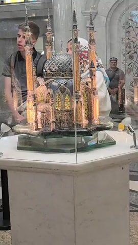 This media file shows a close-up view of an ornate model of a mosque, displayed inside a glass case. The mosque model is highly detailed, featuring multiple minarets, domes, and intricate decorative elements. It appears to be made of metal, possibly gold or brass, with colored glass windows. The model rests on a clear glass platform, which is elevated on a marble pedestal.

In the background, partially visible through the glass, are several people. To the left, a young man with short brown hair, wearing a dark grey t-shirt and a backpack, is looking towards the mosque model. To the right, further back, a person wearing a white hijab with a colorful pattern is also observing the display. Behind them, another person, wearing a dark shirt and a patterned cap, is seated.

The setting appears to be an indoor exhibition or museum, indicated by the polished floors and the lighting. The overall atmosphere is one of quiet observation and appreciation of the detailed craftsmanship of the mosque model. There are no overt signs of specific time of day or weather conditions. The location context provided is Usady, Russia, suggesting this could be a cultural or historical exhibit within that region. No visible text is present in the media file.