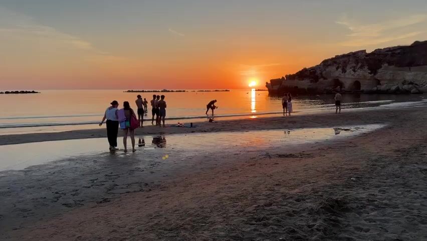 This is a video of a beach scene at sunset. The sun is low on the horizon, casting a warm orange glow across the sky and water. Several people are scattered on the beach and in the shallow water. On the left side of the frame, a woman in a swimsuit and with a pink backpack is taking a photo with her phone. Beside her, a group of people stand together. In the middle ground, a man is bending down in the water. To the right, two people stand together on the beach, and another person stands further down the shore. The water is calm and reflects the colors of the sunset. The beach is sandy, with some wet sand and small pools of water reflecting the sky. A rocky cliff formation is visible on the right side of the frame. The overall atmosphere is peaceful and relaxed, with people enjoying the evening at the beach.