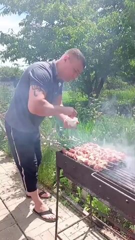 A man is grilling meat skewers over a charcoal grill in an outdoor setting. He is wearing a dark t-shirt, black pants, and flip-flops. The grill is placed on a paved area, with greenery and trees in the background. Smoke is rising from the grill, indicating the meat is cooking. The sun is shining, suggesting it is daytime. The scene appears to be a backyard or garden in a rural or suburban area. The man seems focused on the task of grilling.