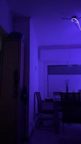 The video shows a dining room at night, illuminated by purple LED strip lights. The lights are attached to the ceiling fan and extend down the wall, creating a dramatic effect. The room contains a dining table with chairs, a cabinet, a vacuum cleaner, and a window with blinds. The overall atmosphere is modern and moody.