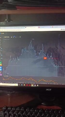 The video displays a computer monitor showing a trading chart. The chart, which appears to be for EURJPY OTC, features a line graph indicating price fluctuations over time. Below the main chart, there is another graph, likely an indicator, with two oscillating lines in yellow and orange. The screen also shows various icons and text related to trading platforms and settings. A keyboard is visible in the foreground. The overall environment suggests a personal workspace focused on financial trading.