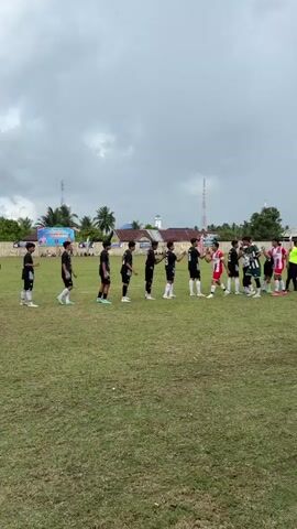 The video shows a group of young soccer players in uniform lined up on a grassy field. Some players are wearing black and white uniforms, while others are wearing red and white striped uniforms. They are shaking hands with each other, seemingly as a pre-game or post-game ritual. There are also a few individuals in referee uniforms present. The background shows a somewhat overcast sky, some buildings, and palm trees, suggesting an outdoor setting, likely in a tropical or subtropical region. The overall atmosphere appears to be one of sportsmanship and friendly competition.