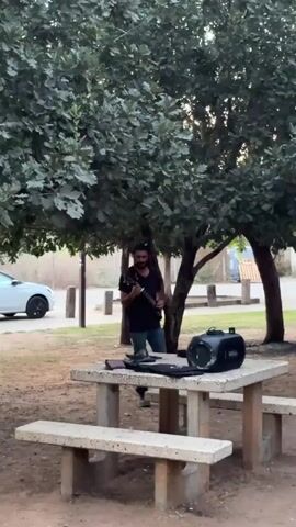 A man is playing an electric guitar in an outdoor setting under a tree. He is wearing a black t-shirt and blue jeans. A portable speaker is placed on a stone picnic table in front of him. The man appears to be performing.