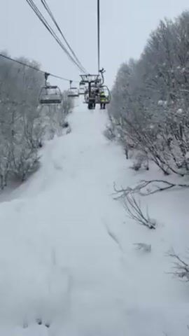 The video shows a ski lift ascending a snow-covered hill.  Two people are visible on one of the chairs, with one wearing a yellow jacket. Trees covered in snow line the sides of the hill.  The scene is overcast.