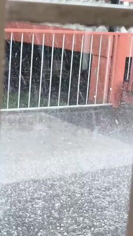 The video captures an outdoor scene in Quito, Ecuador, during what appears to be a heavy hail storm. No people are visible. The ground, possibly a patio or driveway, is covered with numerous small, white hailstones that are actively bouncing and accumulating. In the background, a white fence stands in front of an orange-colored wall, possibly part of a building. The hailstones are falling densely, creating a thick, white layer on the ground. No discernible text is visible in the frame.