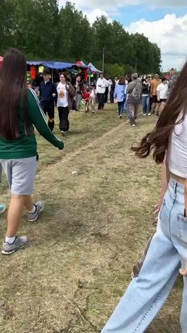 This video shows an outdoor event or market on a cloudy day. There are many people walking around, some near tents and stalls, others in a large open grassy field. In the foreground, a person with long dark hair walks away from the camera, wearing a green and white jacket and grey shorts. Further into the scene, two people, likely a mother and child, walk towards a table displaying balloons and other items. In the background, there are trees, hills, and a large cell tower. The ground is covered in dry grass, with some areas appearing worn down by foot traffic. The overall atmosphere seems to be of a lively gathering.