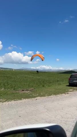 A person sitting in a paragliding seat in a grassy field is holding onto the lines that are attached to an orange and black striped paragliding wing. The person is silhouetted and wearing a dark helmet. The paragliding wing is inflated in the sky. The ground is green grass, and in the distance, there are mountains and a few white clouds. A gray car is parked in the grass on the right of the frame. The camera is at ground level and is likely filming from a car. The sky is blue and clear.