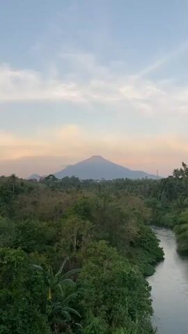 The video begins with a wide shot of a river flowing through a lush, green landscape. In the distance, a large mountain can be seen under a soft, pastel-colored sky. The camera then pans down to follow the river, revealing the riverbank lined with dense vegetation. As the camera moves closer, a person is seen standing in the shallow water, appearing to be washing clothes or performing some other activity. They are partially obscured by the water and vegetation, but they are wearing dark shorts and a blue top. There are several items, possibly laundry baskets or bags, near the person on the riverbank. The water is murky and flowing. The overall scene is serene and natural.