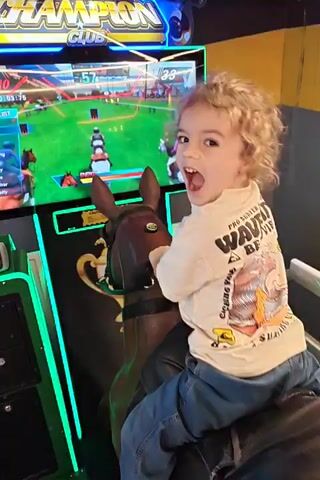 A young child is riding a horse-shaped video game in an arcade. The game is “Derby Champion Club”, a horse racing simulation with a screen showing a race in progress. The child is wearing a white t-shirt with a “Wave Beach” graphic and blue jeans. They are holding onto the reins of the horse and are engaged with the game. The arcade has yellow and grey walls.