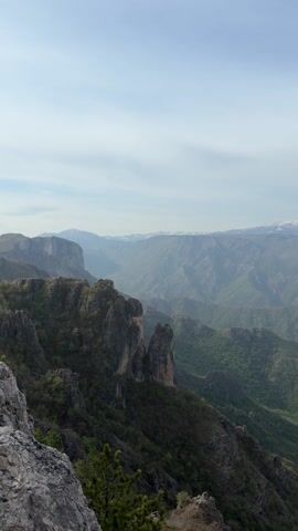 The video shows a view of green, mountainous terrain under a bright sky. In the foreground are rocky, jagged cliffs with vegetation. Beyond these, a landscape of layered mountain ranges stretches into the distance, with peaks capped with snow visible in the far background. The sky is a pale blue with a light haze or thin clouds. The camera pans slowly to the right, showcasing the expanse of the mountain range.