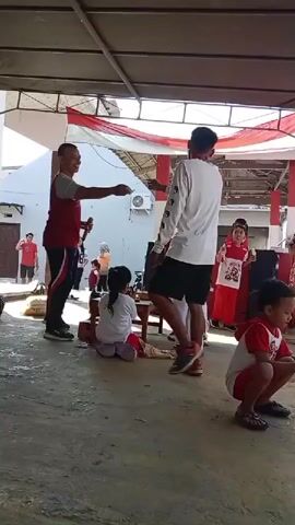 The video shows an outdoor event with several children and adults. In the foreground, a man in a white shirt and black shorts stands facing a group of children. He appears to be leading an activity, possibly a game, as he holds a microphone. Several children, dressed in red and white, are gathered around. Some are standing, while others are sitting or crouching on the ground. One girl in a red shirt and white pants is holding a white object. In the background, there is a white building with red and white striped fabric draped across the ceiling of an open-air structure. There are also some black speakers and a small wooden table visible. The ground is paved with concrete. The lighting suggests it is daytime.