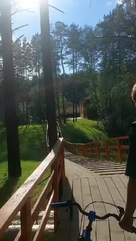 This media file captures a first-person perspective of riding a bicycle across a wooden bridge. The bridge is lined with a wooden railing, painted a warm orange color. Beyond the railing, a lush green grassy slope is visible, dotted with tall pine trees that create dappled shade. In the distance, a building with a red roof is nestled amongst the trees.

The activity is a leisurely bike ride. Two individuals can be seen walking on the bridge ahead of the cyclist. The time of day appears to be daytime, with bright sunlight casting long shadows, suggesting it might be late morning or early afternoon. The weather seems pleasant, indicated by the clear sky and the verdant landscape. The presence of trees, a bridge, and grassy areas points to a natural, possibly park-like or recreational setting. The overall mood conveyed is one of peaceful enjoyment of the outdoors. There is no visible text in the media file.