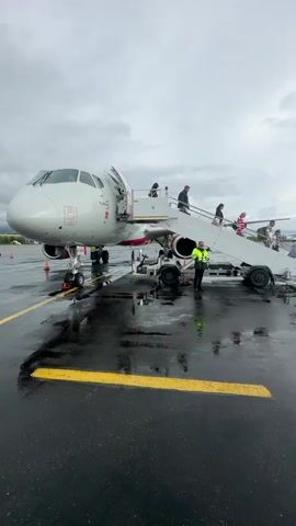 A person in a bright green vest stands on the tarmac by a set of mobile stairs that lead up to an airplane door. Several people are walking up the stairs into the plane. The tarmac is wet, with reflections on the surface, suggesting it might be raining or has recently rained. The sky is overcast. The front of the airplane is visible, as are its engines and landing gear. Yellow lines are painted on the tarmac.