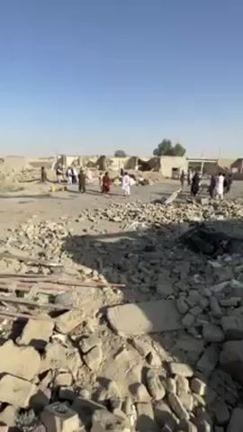 The video shows a devastated area filled with rubble and debris, likely the aftermath of a bombing or other destructive event. The ground is covered in broken bricks, concrete chunks, and twisted metal. In the background, damaged buildings stand with crumbling walls. Several people are visible in the distance, some wearing traditional clothing. They appear to be surveying the scene or searching through the wreckage. The sky is clear and blue, and the overall atmosphere is somber and chaotic.
