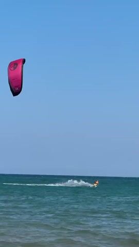 A pink kite is visible in the upper left corner of the frame against a clear blue sky. Below the kite, on the surface of the ocean, a person is kitesurfing. The surfer is wearing an orange life vest and riding a board that is kicking up a spray of water. Another kite, this one colored green and white, is visible closer to the surfer. The ocean is a deep blue with moderate waves.