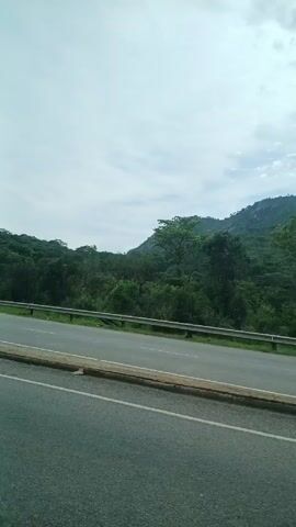 The video shows a view from a car window, looking out at a two-lane highway winding through a wooded area with a low mountain in the background. A large semi-truck is visible traveling in the opposite direction. The sky is overcast.