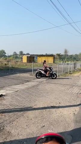 A person rides a white and black scooter down a gravel path. The person is wearing a brown top and dark shorts. They are looking forward. Behind them is a chain-link fence and a yellow building. The sky is clear and blue.