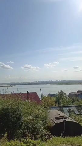 A time-lapse video shows a bright sunny day with a clear blue sky and scattered white clouds over a wide body of water. In the foreground, lush green bushes and trees partially obscure a red-tiled roof of a house. Further to the right, a large metal barrel is visible, and to the left, a greenhouse with a blue barrel next to it. The water stretches to the horizon, where a faint cityscape can be seen in the distance. The sun is high in the sky, casting bright light on the scene. The camera slowly pans from right to left, revealing more of the greenhouse and surrounding vegetation.