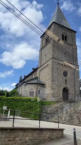 The video shows a stone church with a tall bell tower against a blue sky with white clouds. The church has a sloping roof and a pointed spire. There are power lines running across the top of the frame. A set of stone stairs leads up to the entrance of the church. The church is partially covered in ivy on the left side. A yellow banner with German text and cartoon children is attached to the fence in front of the church. There is a traffic sign with a picture of children walking near the church entrance. The camera pans slightly upward, showing the bell tower in more detail. A fence encloses the church grounds.