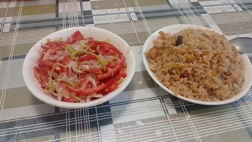Two white bowls of food are presented on a checkered tablecloth. The bowl on the left contains a tomato and onion salad, with sliced tomatoes and thinly sliced white onions, possibly with some green herbs. The bowl on the right contains a pilaf, a rice dish mixed with chickpeas, what appears to be small pieces of meat, and some dried fruit. A spoon is partially visible to the right of the pilaf bowl. The setting appears to be a dining table, likely indoors, given the lighting and the tablecloth. There is no visible text in the image. The context provided indicates the location as Mailuu-Suu City, Kyrgyzstan.