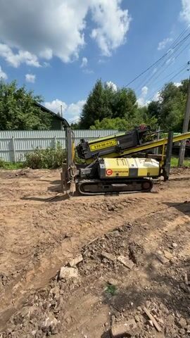 A horizontal directional drilling machine, yellow and black in color, is positioned on a dirt surface. It has a long boom extending from its rear. The machine is situated next to a shallow trench that has been dug into the ground. A white fence and trees are visible in the background, with a blue sky and white clouds above. The area appears to be under construction or undergoing some form of groundwork.