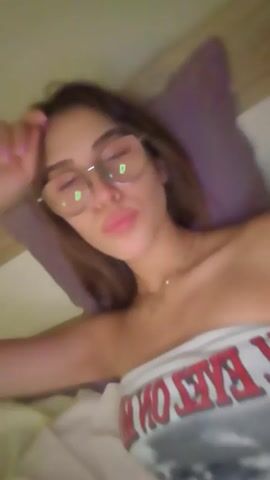 A woman with long brown hair and glasses is lying in bed. She is wearing a strapless top with red lettering. She is looking at the camera and puckering her lips. The background is a pillow and a sheet.