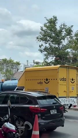 This media file captures a street scene in Chiang Mai, Thailand. The primary focus is on a utility vehicle, specifically a water truck, actively spraying water. A person, dressed in a green shirt and a hat, is visible operating the spraying mechanism from the back of the truck. The truck itself is a light blue color with a large tank.

In the foreground, a black SUV is parked, and its rear is clearly visible. Next to it, parts of a pink and white scooter are also in view. A bright orange traffic cone stands near the SUV's rear wheel. Behind the water truck, a bright yellow truck with "uda express" branding is visible, featuring a smiley face logo and QR codes.

The setting appears to be an urban environment with buildings and trees lining the street. The weather is overcast, with a cloudy sky suggesting it might be during the day, perhaps late morning or afternoon. The activity is clearly related to street cleaning or perhaps watering the roadside greenery, indicated by the water being sprayed. The general mood is neutral, focusing on the everyday activity. Visible text includes "uda express" on the yellow truck and Thai characters on the license plate of the black SUV, which reads "จจ 948". There is also Thai text on a banner on the water truck.
