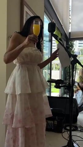 The video opens with a woman in a white and pink strapless dress holding a glass of orange juice and a piece of paper near a microphone. She appears to be speaking or about to speak. The camera then pans to a group of women sitting at a table in a brightly lit room with large windows. The women are smiling and appear to be engaged in a conversation or event. The room has a modern aesthetic with plants and architectural elements visible through the windows. A microphone and sound equipment are visible in the foreground, suggesting a presentation or performance is taking place.