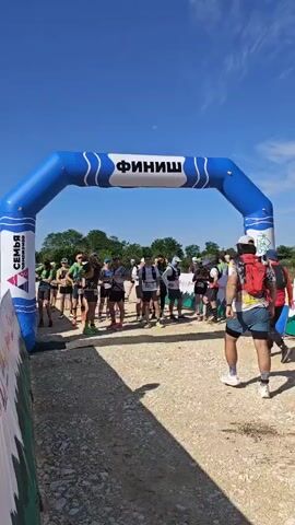 A vertical shot shows a group of people standing under a blue inflatable arch with "ФИНИШ" (FINISH) written on it. They are dressed in athletic attire and appear to be waiting for something to begin. The man closest to the camera is wearing a white cap, a patterned shirt, black shorts, and a red backpack, and he's looking towards the right side of the frame. The other people are looking around or talking to each other. The background is a bright blue sky with some trees. The ground is dirt and gravel.