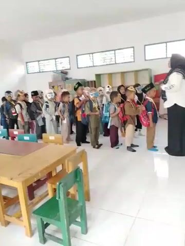 In a brightly lit classroom, a line of approximately 20 young children, all wearing backpacks, are standing in an orderly queue. Most are wearing dark hats and decorative masks. An adult woman, wearing a white blouse and a black skirt, stands facing the line, seemingly instructing them. Tables and chairs are scattered around the room, indicating a learning environment. Several windows let natural light into the space. The overall atmosphere appears organized and potentially educational.