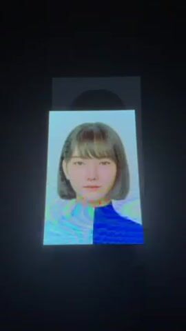 The video shows a photo booth display with a portrait of a young woman with short, straight hair and bangs. Her clothing is half light blue and half dark blue. The background is a dark grey. An opening for dispensing the photo is visible above the displayed image. There is some indistinct ambient sound.