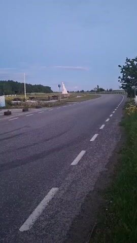 A silver car drives along a winding asphalt road. The car is in motion and blurred due to the camera's shutter speed. The road has white dashed lines painted on the right side. On the left side of the road, there is a sculpture or statue. The sky is a clear, pale blue, and the sun appears to be setting or has recently set, casting a dim light on the scene. Green grass and yellow flowers grow along the right edge of the road. A tree with dark green leaves is visible on the far right. The background consists of a flat landscape with some distant trees.