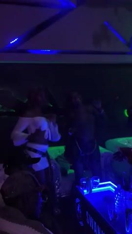 A group of young women are dancing and socializing in a dimly lit nightclub. The atmosphere is vibrant with flashing blue and purple lights, and music can be heard. The women are dressed in stylish club attire, and they appear to be enjoying themselves. One woman with pink hair is at the center of the scene, dancing energetically. Another woman in a white top and skirt is also dancing. The background shows a bar area with bottles and glasses, illuminated by neon blue lights. The overall impression is of a lively and fun night out.