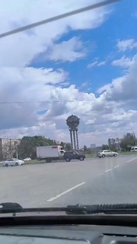 The video shows a view from inside a vehicle traveling through an urban intersection on a cloudy day. In the foreground, part of the car's dashboard and windshield wipers are visible. Beyond the windshield, several cars are seen: a white truck with a white box on the back, a dark SUV, a silver sedan, and another white sedan. A large monument with a star on top, supported by three tall columns, is prominently displayed in the background, situated on a grassy roundabout. Apartment buildings and trees are visible behind the monument and to the sides of the intersection. The sky is partly cloudy with some darker clouds suggesting potential rain. There is no discernible interaction between the vehicles, and no individuals are clearly visible.