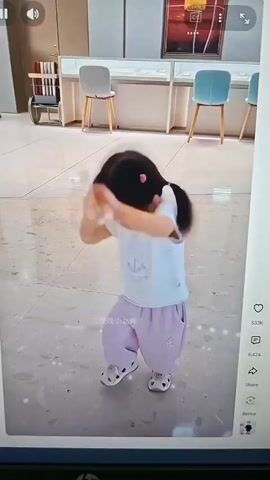 A young East Asian girl with black hair in pigtails, wearing a white t-shirt with a cartoon mouse print and light purple pants, stands on a tiled floor. She is in motion, with her arms extended and hands open. Her head is tilted back slightly, and her eyes are closed. The background features display cases with fabric coverings, chairs, and a wheeled cart with a colorful back. The lighting is bright, and the scene appears to be indoors, possibly a retail environment. The video is timestamped from 00:00 to 00:05.