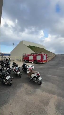 The video shows a red fire truck parked beside a building on a cloudy day. Several motor scooters are parked to the left of the truck. A paved road leads uphill to the right of the fire truck. A low stone wall is on the right side of the road, with a grassy hillside on the left. In the distance, a line of trees can be seen at the top of the hill.