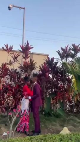 A young African-American couple is embracing in front of a tall hedge of red-leafed plants. The woman is wearing a red top and a long, floral patterned skirt. The man is wearing a dark purple suit. They are standing on grass in front of a building. There is a tall light post with a security camera atop it nearby.